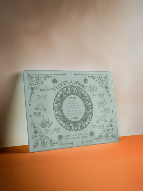 A rectangular sage-tinted glass cutting board featuring an ornate design of magical herbs and spices. Text reads "Love's Alchemy" with detailed illustrations of vanilla, rose, cardamom, star anise, basil, and cinnamon arranged around a central medallion containing a love spell verse.